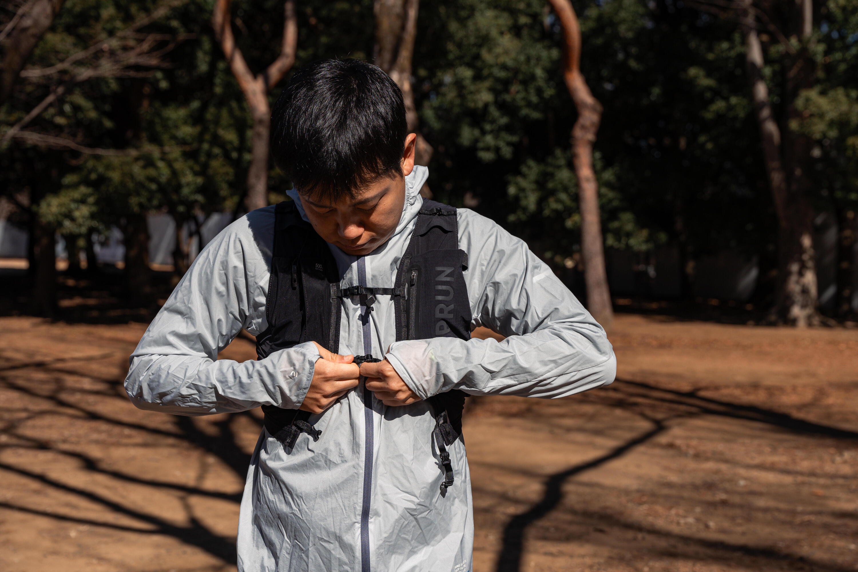 Man fastening a grey running jacket in a park, with a black running vest underneath