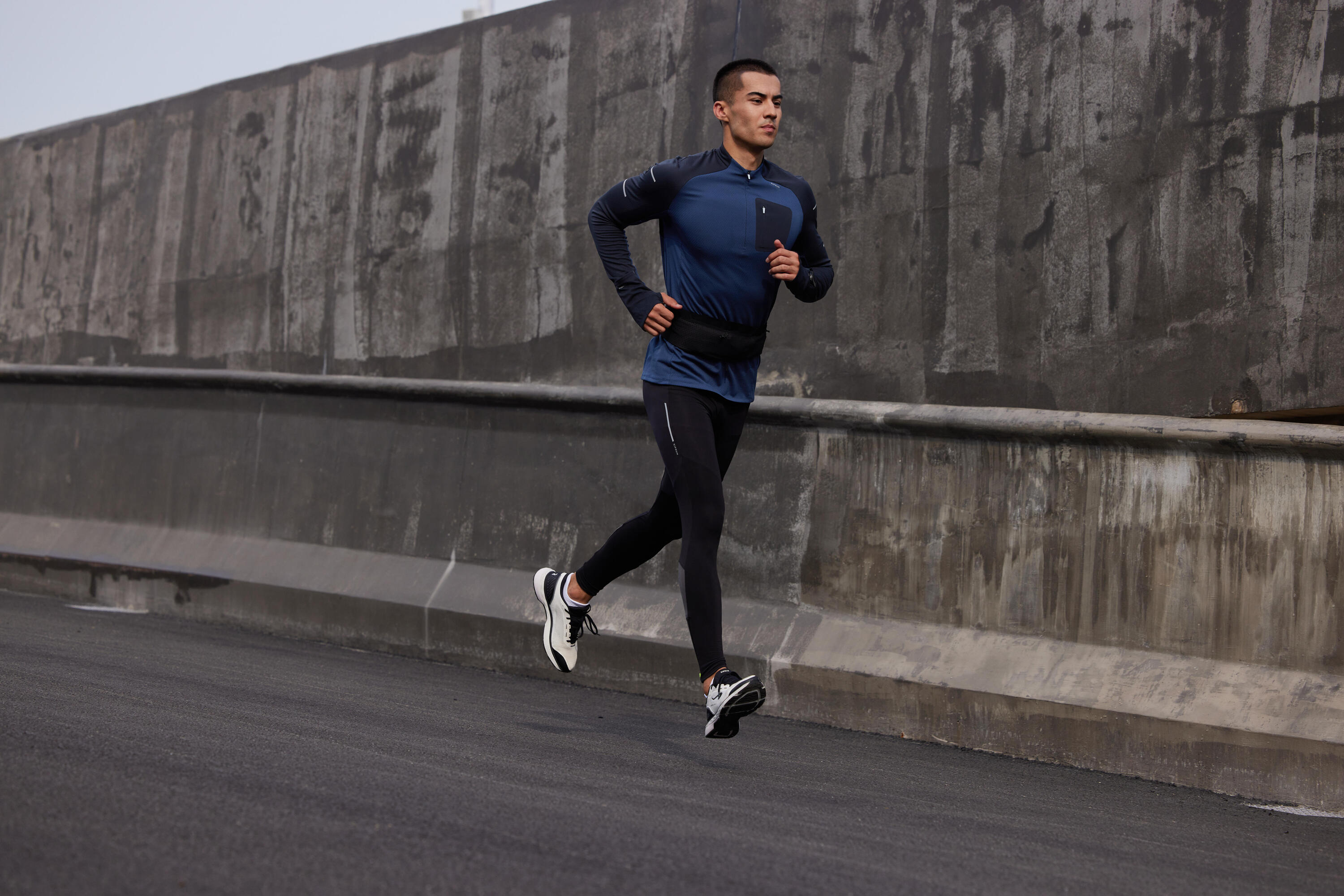 Man running on an urban road wearing a black running belt over a blue long-sleeve top and tights
