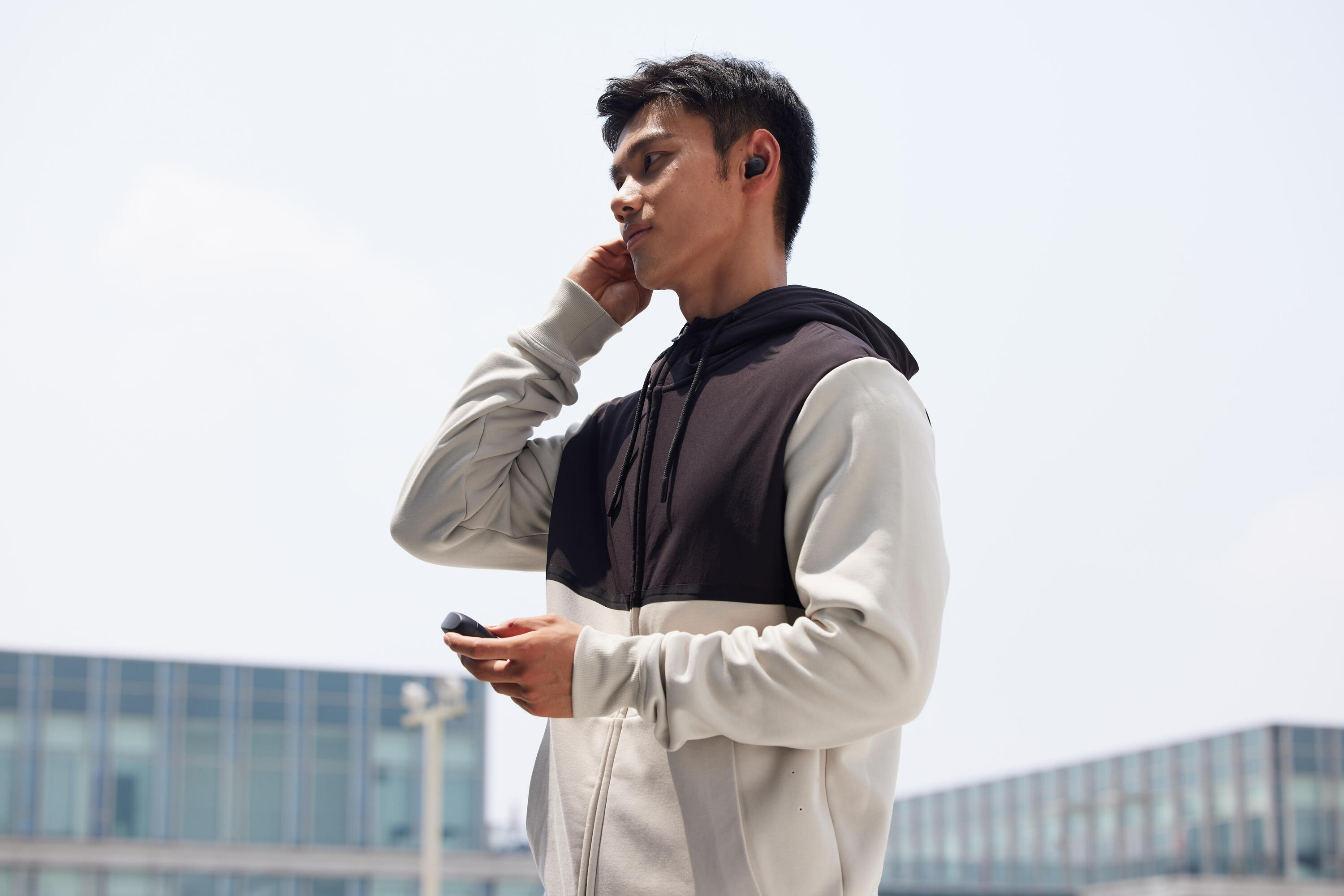 Man adjusting a wireless earbud outdoors, holding the charging case, in a black and grey hoodie