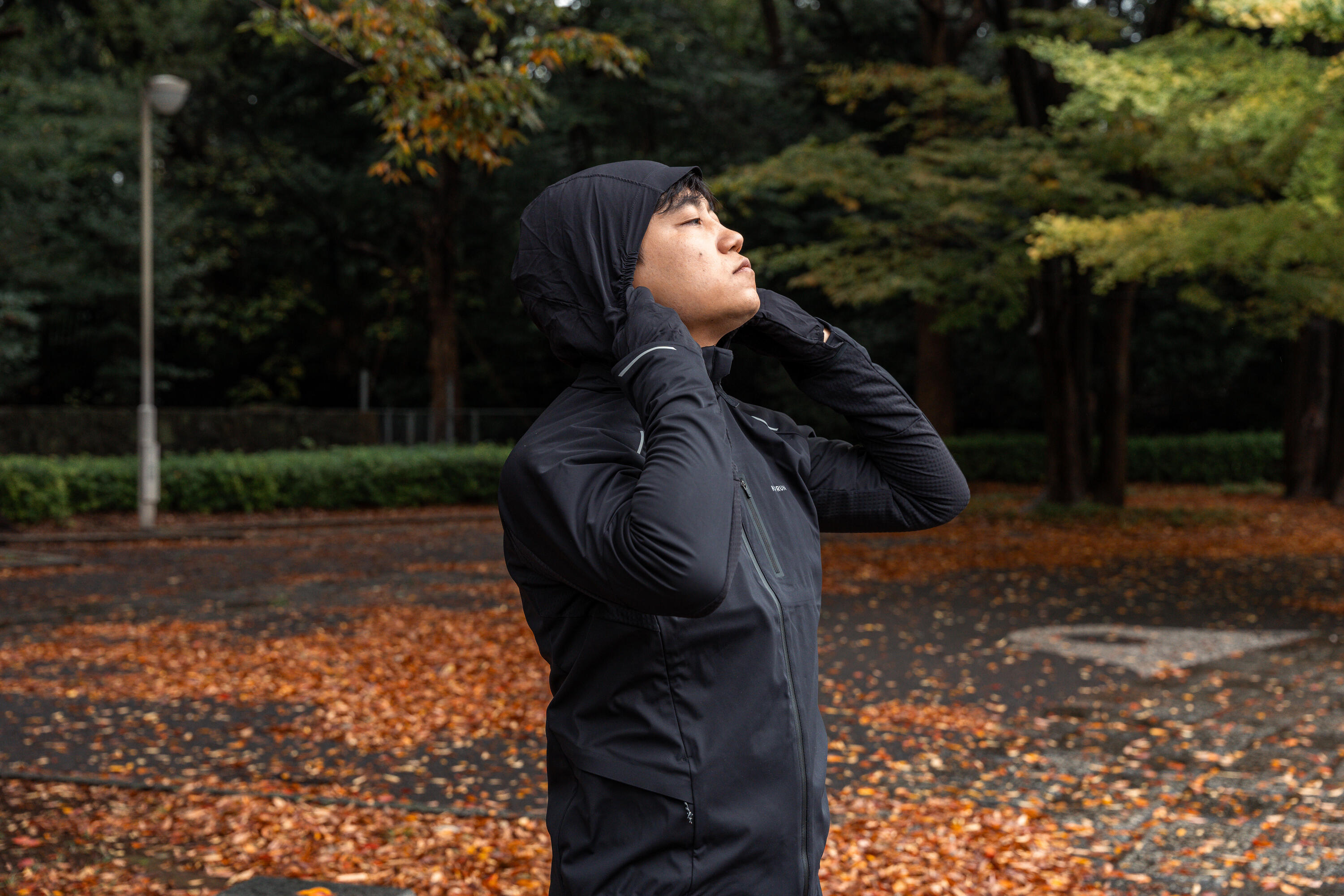 Man in a black hooded running jacket pulling up the hood in an autumn park
