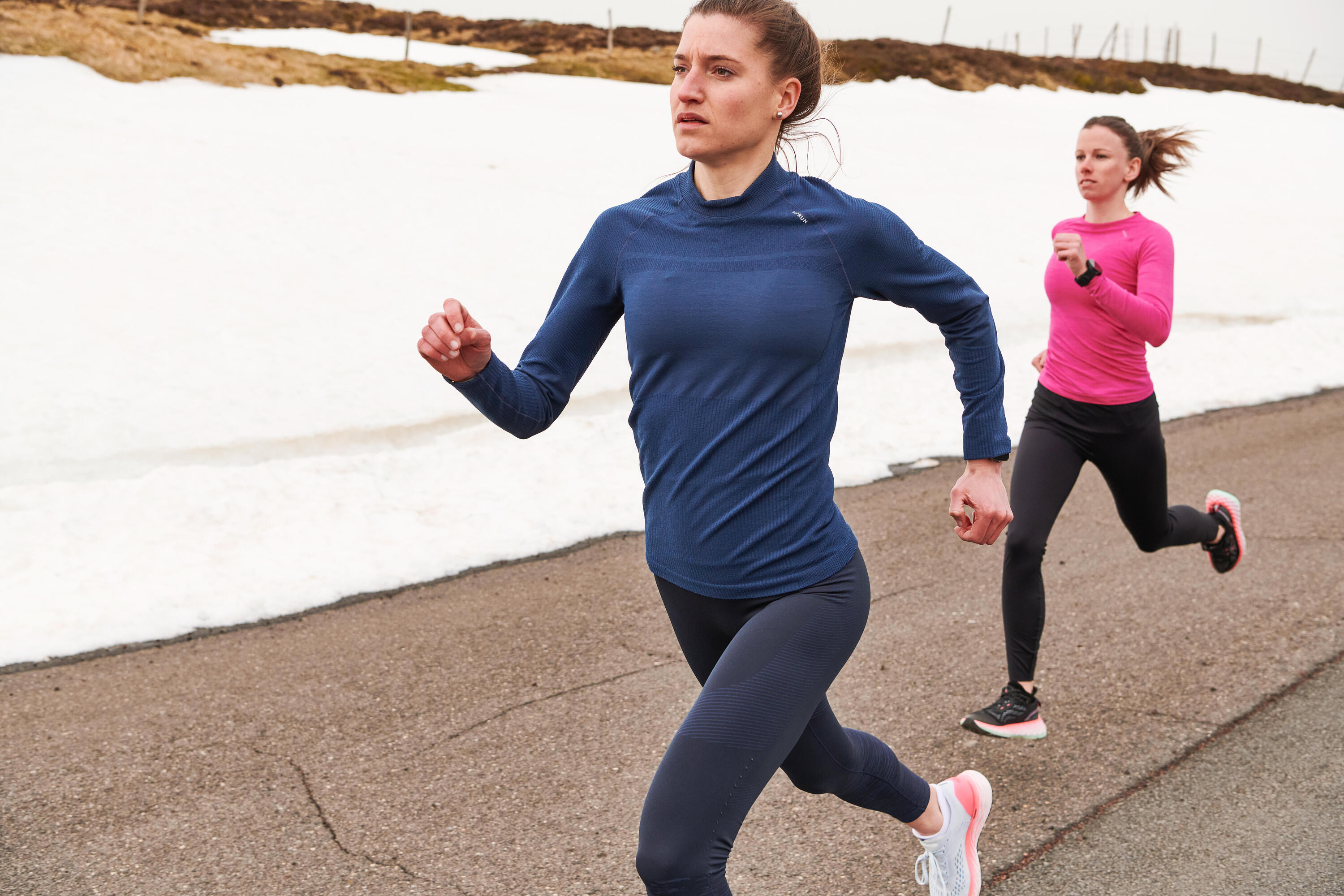 Two women running on a snowy mountain road in long-sleeve tops and running leggings