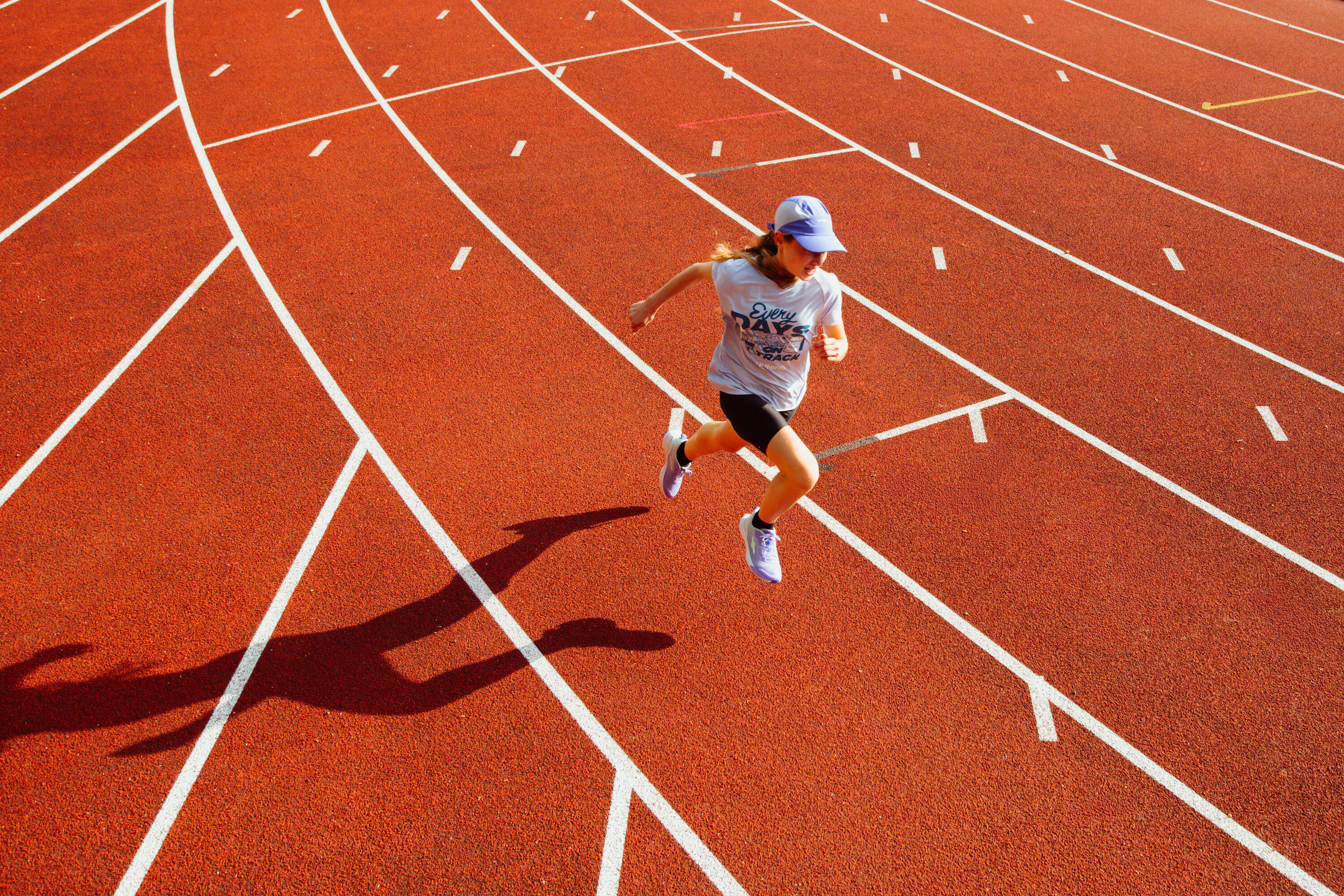 Girl sprinting on a red athletics track seen from above, casting a long shadow, in a white top and cap