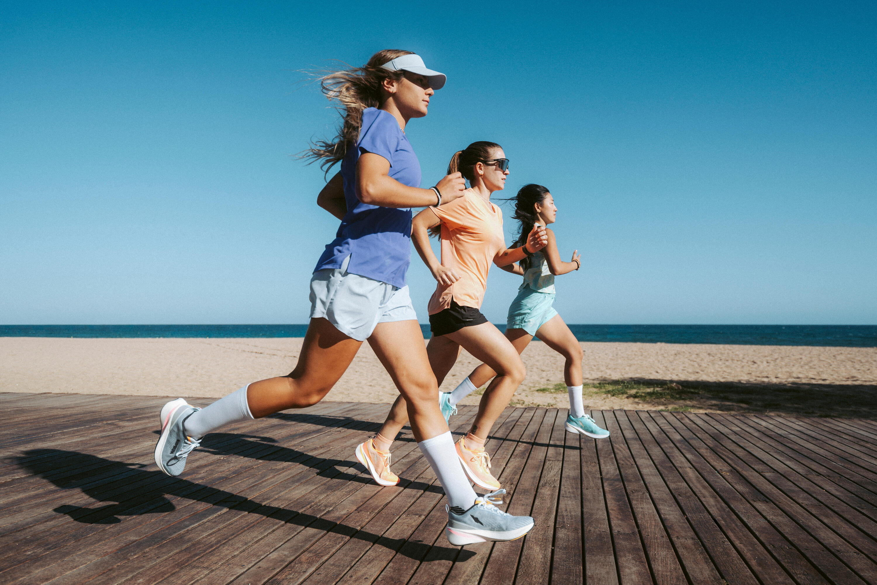 Three women running on a beachfront boardwalk in colourful tops and shorts