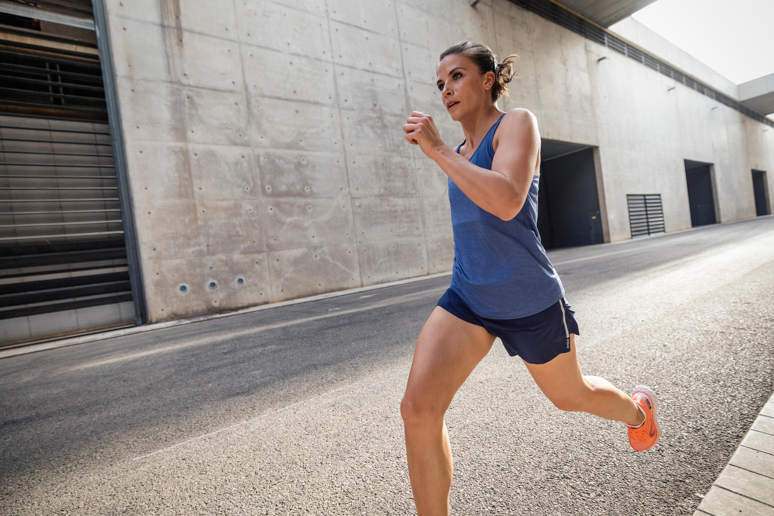 Woman running along an urban concrete street in a blue vest, navy shorts and orange running shoes