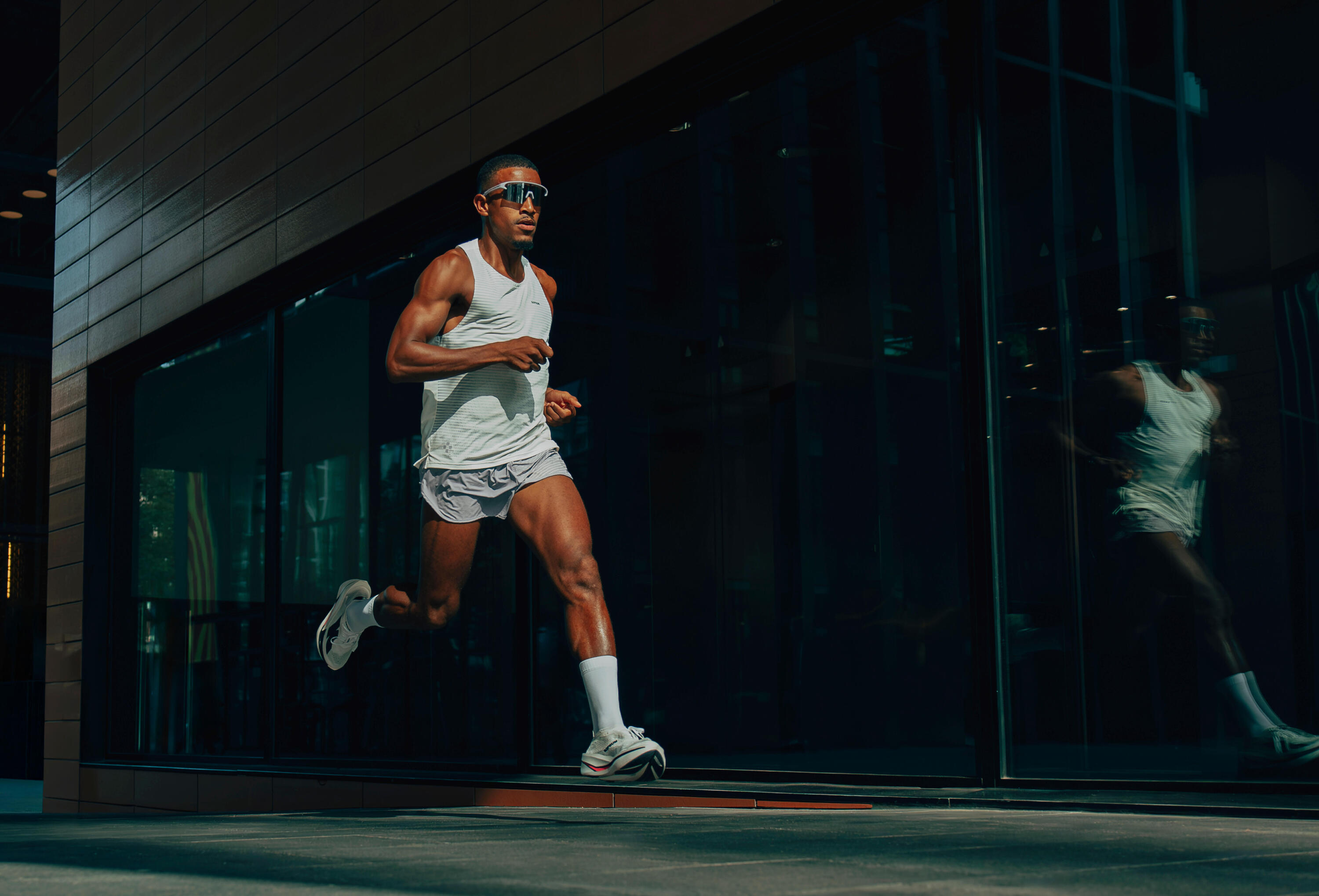 Man running past a dark glass building in a white running tank top and grey shorts
