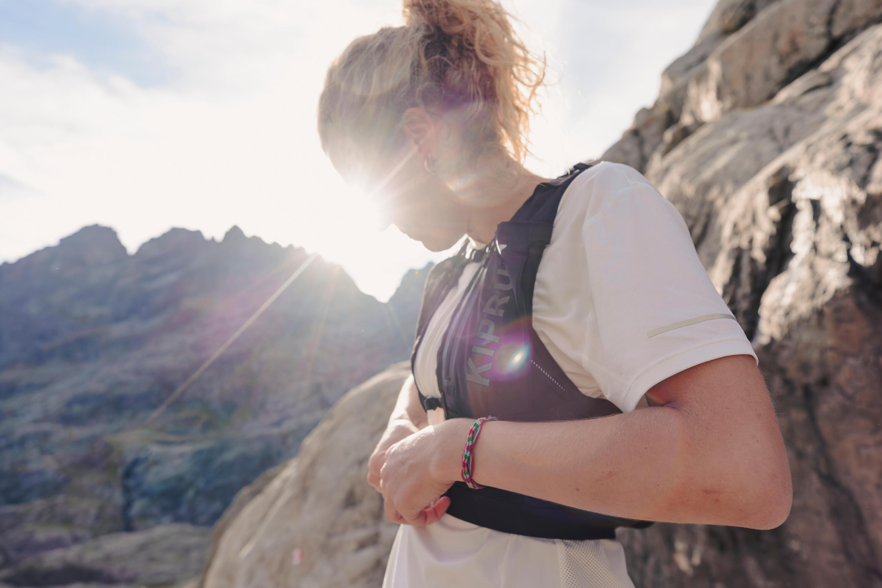 Woman adjusting her hydration vest on a mountain trail at sunrise, wearing a white top