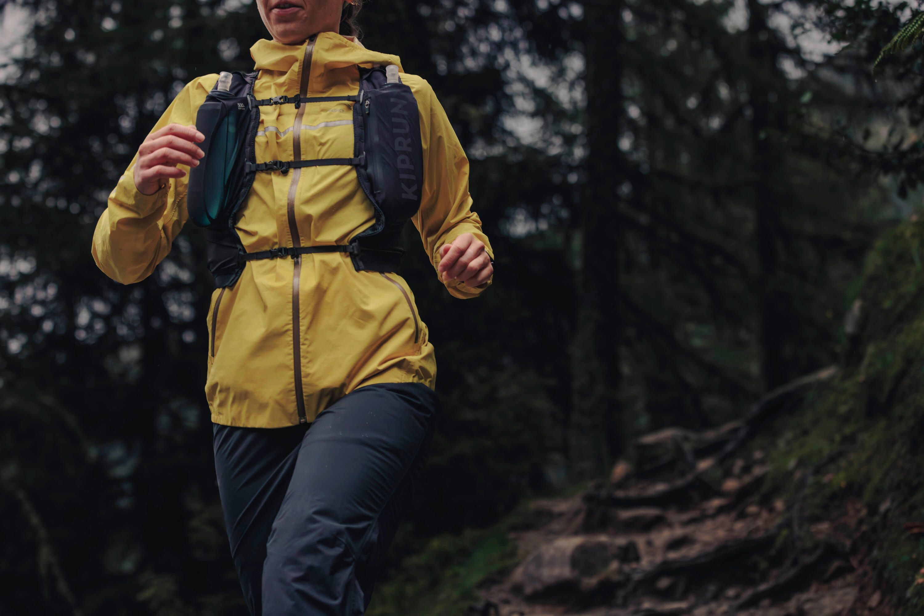 Trail runner in a yellow jacket and black Kiprun hydration vest with soft flasks, running through a forest
