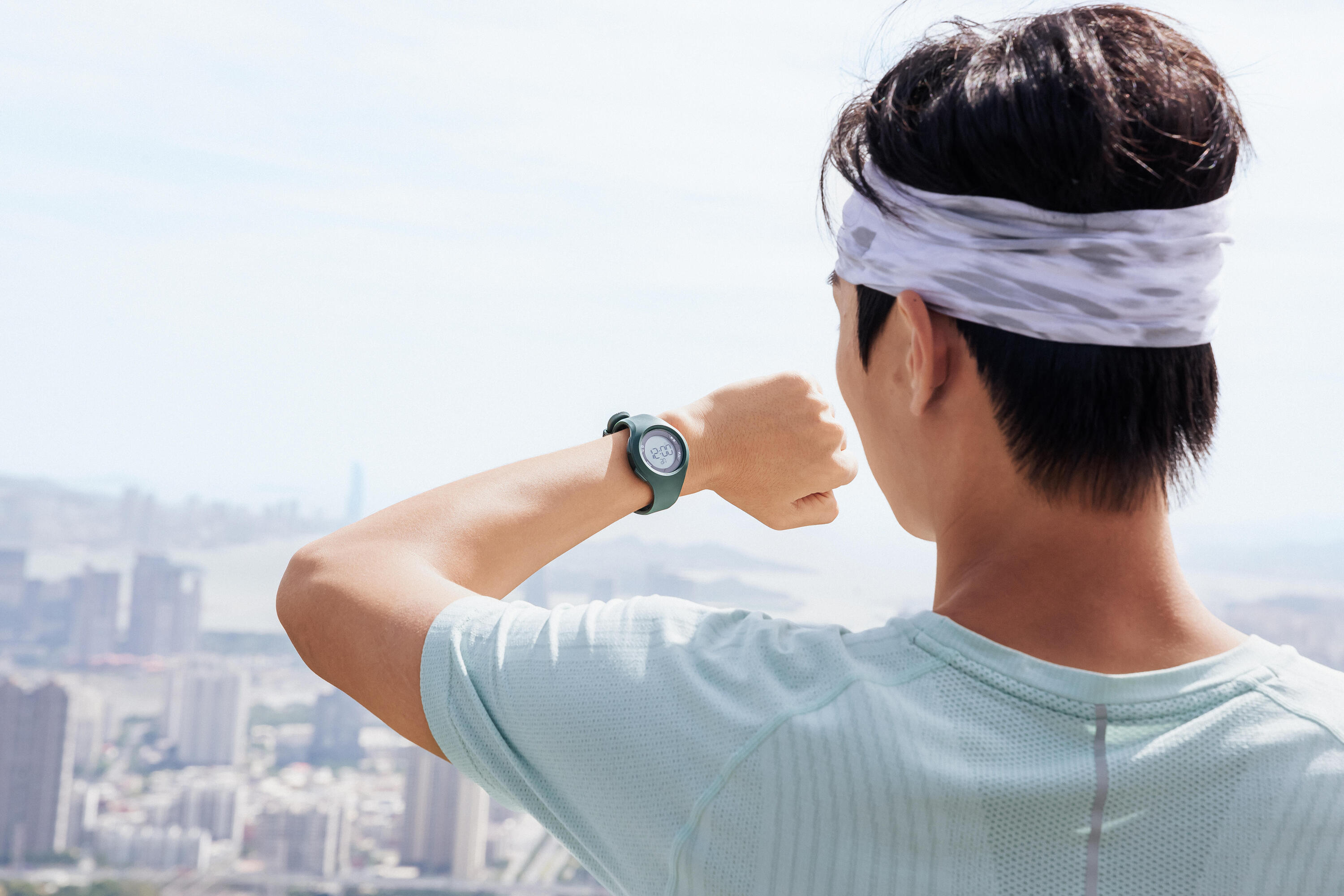 Man checking his sports watch while looking out over a city skyline, wearing a headband and mint t-shirt